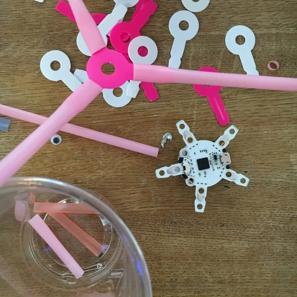Parts for the Dancing Robot workshop on a table: a small circuit board, pink drinking straws and white plastic connectors.