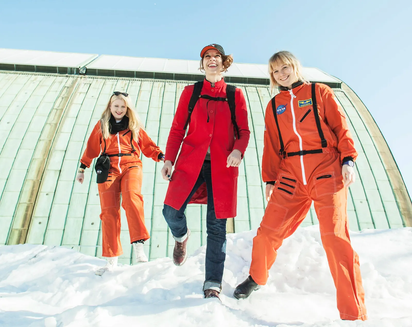 Three women in orange and red space suits walking through snow in front of a domed building.