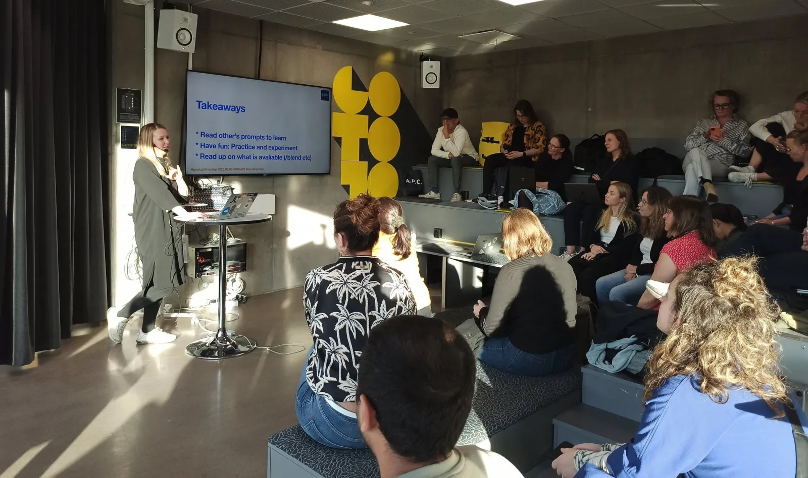 A speaker presents a "Takeaways" slide to a seated audience, in a room with large yellow letters on the wall.