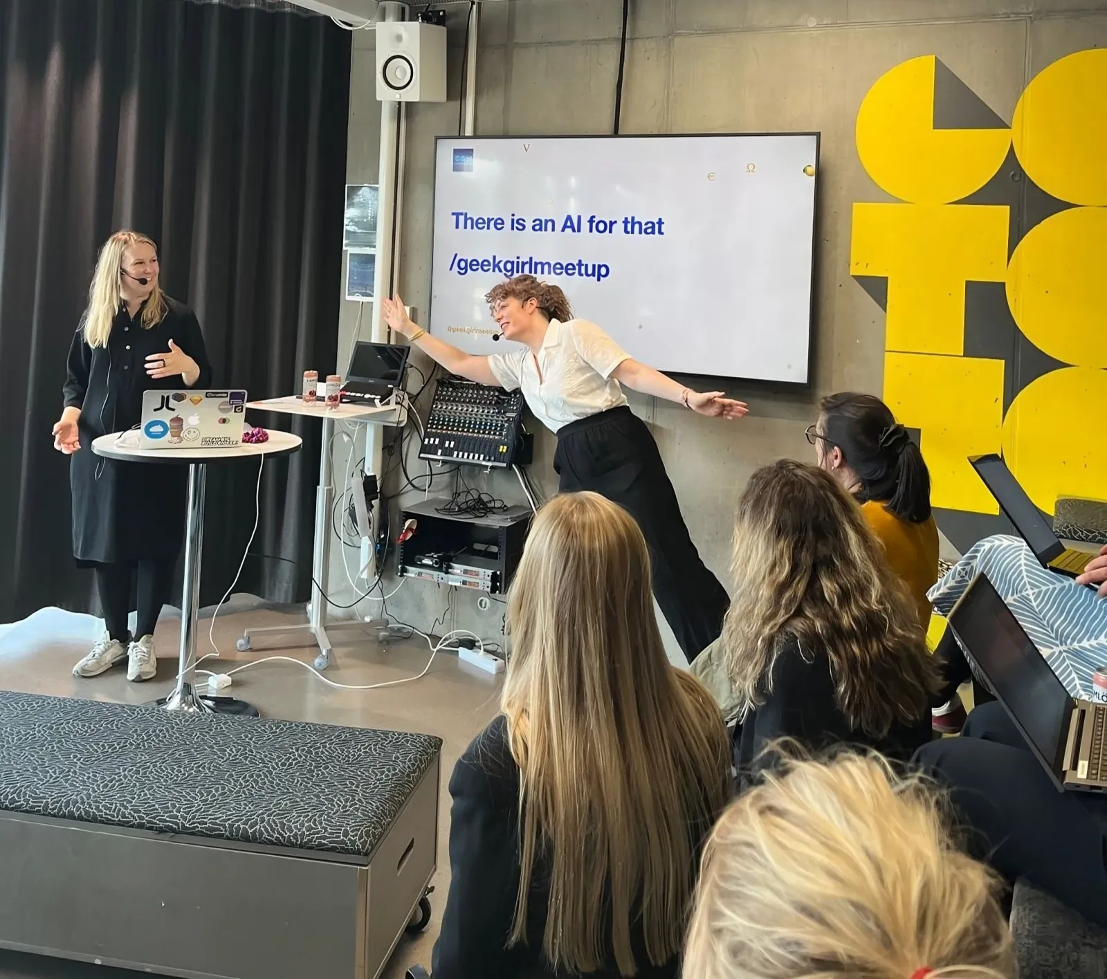 Two presenters in front of a screen reading "There is an AI for that", with the audience in the foreground.
